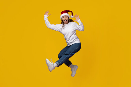 A woman jumps with excitement while wearing a Santa hat and casual clothing against a vibrant yellow backdrop. Her joyful expression captures the festive spirit of the holiday season. - Powered by Adobe