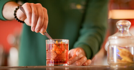 Obraz premium Garnishing cocktail. Alcoholic mix. Barman preparing refreshing drink with ice cube. Cold negroni served with orange fruit zest in glass.