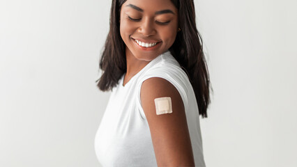 Healthcare And Inoculation Concept. Smiling Black Female Patient Showing And Looking At Vaccinated Arm With Plaster On Shoulder After Coronavirus Vaccination. Studio Banner Panorama. Antiviral Vaccine