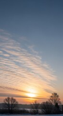 Fototapeta premium Sunrise over snow covered field with bare trees and cloud formations