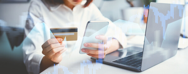 A focused young businesswoman in glasses sits at her office desk, analyzing problems with a banking app on her smartphone while holding a credit card, indicating a financial concern during her work.