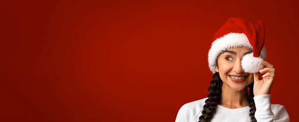A young woman wearing a red Santa hat playfully adjusts the hat's pom-pom while smiling. The bright red background adds to the festive atmosphere, capturing the spirit of holiday joy and celebration.