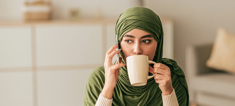 A young woman wearing a green hijab drinks from a white mug while engaged in a phone conversation. She sits in a cozy home setting with soft lighting and minimal decor. - Powered by Adobe