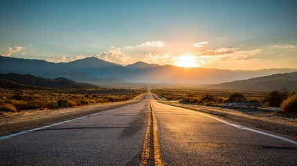 Fototapeta premium A long road stretches into the distance surrounded by hills and mountains. The sun sets on the horizon casting light on the landscape. The sky shows shades of orange and blue.