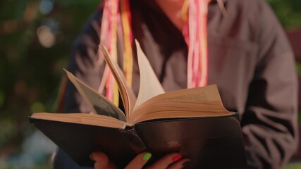 person reading open novel under trees on bench, soft shade and warm highlights, fingers holding spine and flipping pages, casual jacket and colorful hair strands, peaceful park study scene