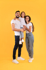 A cheerful family stands together in casual clothing. They display joy and love, with a child playfully lifted between parents in a bright yellow setting.