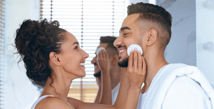 A couple enjoys a light-hearted moment in the bathroom, applying skincare products. They smile at each other while standing in front of a mirror, creating a warm and playful atmosphere. - Powered by Adobe