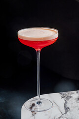 A vibrant red cocktail in a tall glass, topped with frothy foam on a marble surface