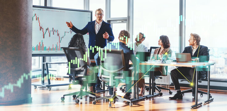 A businessman in a suit stands in front of a whiteboard displaying a financial chart, presenting to a small group of colleagues seated around a table in a modern office with large windows.