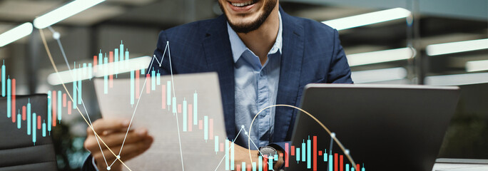 A happy millennial man in a suit sits at his worktable in a modern office, reading a marketing report.