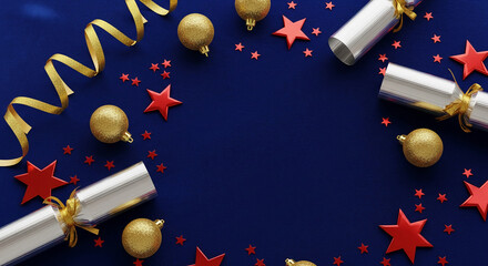 Overhead view of silver cracker, gold ornament and ribbon, and red star on dark blue surface, suggesting festive celebration, party or Christmas theme