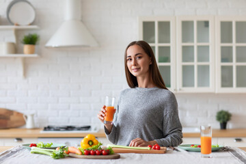 Woman in gray sweater is holding glass of orange juice and smiling at the camera in kitchen with white brick