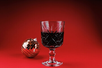 Crystal glass filled with red wine next to a reflective Christmas ornament on red background