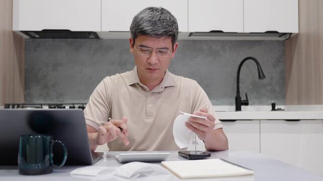 Front view of focused entrepreneur in glasses reviewing bills and calculating monthly expenses at home. Budget planning, financial management and cost control in a modern kitchen environment.