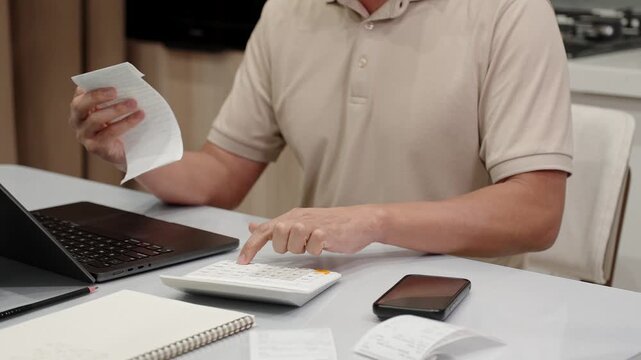 Unrecognized male person reviewing receipts and entering numbers into calculator while managing monthly expenses at home. Financial planning and budgeting workflow.