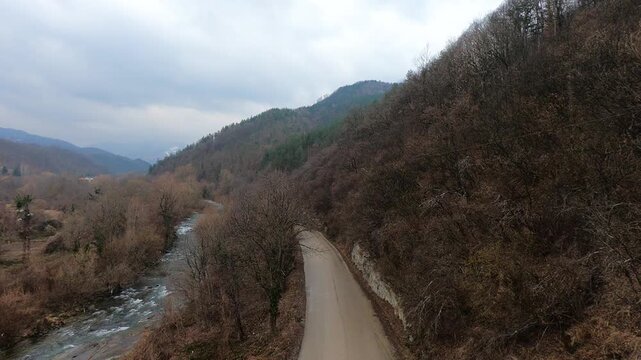 Vertical drone shot of a car driving on a narrow road to Troyan Monastery, flanked by steep forested slopes, a river on the left, under cloudy gray skies, BG, EU, 02 March 2019