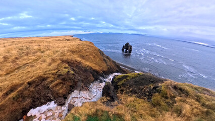 A high-angle view looks out over a sweeping black sand coastline and the blue waters of a bay in northwest Iceland, where a prominent basalt sea stack stands near the shore under a wide sky