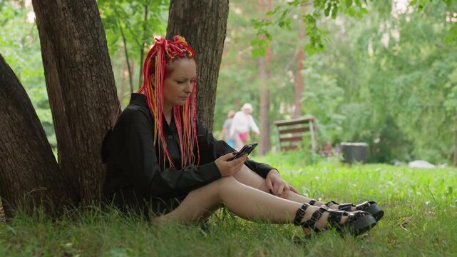 wide shot caucasian student under tree scrolling phone, leaning against trunk with sandals and jacket, blurred passersby in distance, solitude and digital detachment in green park