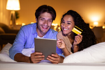 A couple relaxes in their modern bedroom, using a tablet and credit card for online shopping. They happily engage in e-commerce while lying on the bed in a cozy atmosphere.