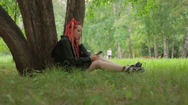 peaceful female rests beneath tree watching others, tranquil woman with red dreadlocks observes moving jogger in park, gentle woman with quiet gaze remains seated as jogger passes by quietly in park