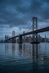 Fototapeta premium Iconic illuminated suspension bridge spanning a wide bay with a modern city skyline of skyscrapers reflecting in the water under a dramatic, cloudy evening sky