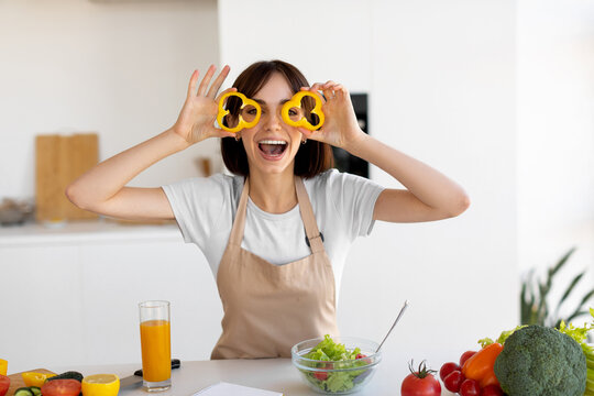 A woman smiles brightly in her kitchen, holding yellow bell pepper slices to her eyes. She is surrounded by fresh vegetables while preparing a healthy salad and enjoying a glass of juice. - Powered by Adobe