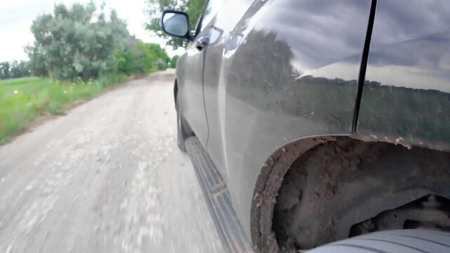 A car moves along a bumpy, unpaved road surrounded by trees. Dust and dirt cover the vehicle, showing the struggle of driving through poor conditions.