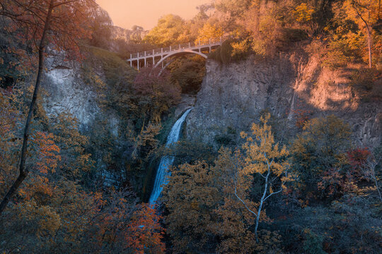 Waterfall streaming down a rocky cliff, surrounded by autumn trees and lush vegetation with a white arch bridge above in Tbilisi botanical garden