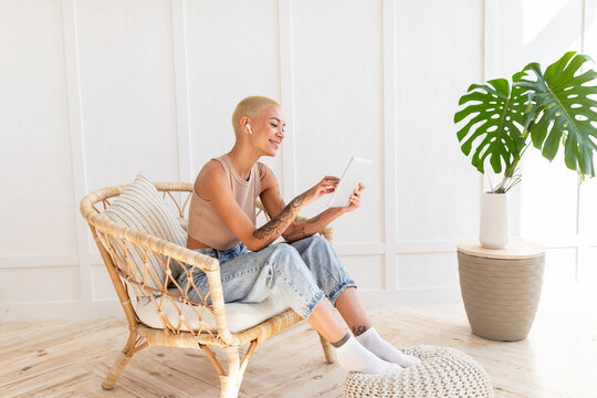Lady using digital tablet and wireless earphones, resting in wicker chair over white wall, copy space. Happy woman watching videos or using application on pad