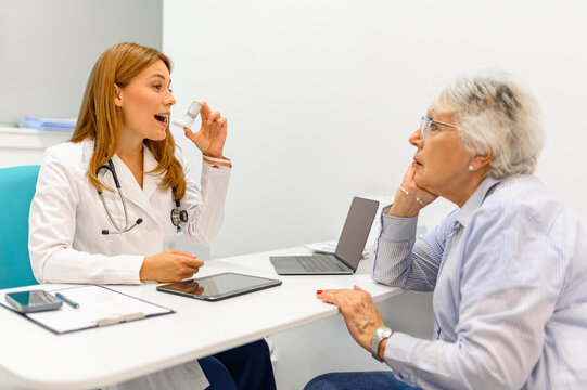 Female doctor with laptop and tablet on desk guiding senior woman how to use asthma pump during routine consultation at clinic - Powered by Adobe