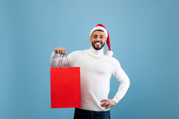 A joyful man wearing a white sweater and Santa hat holds a bright red shopping bag. He stands in...