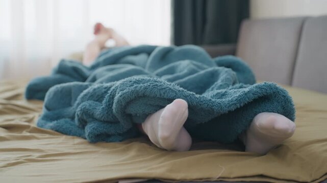 Woman Traveler Feet Peeking From Teal Blanket On Sofa, Striped Socks Visible Against Olive Bedding And Soft Sunlight Playful Cozy Rest Scene Suggesting Journey Pause And Home Recovery.