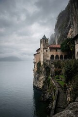 Naklejka premium Ancient stone monastery with a bell tower built into a sheer cliff face overlooking a calm lake on a moody, overcast day with dramatic clouds and fog