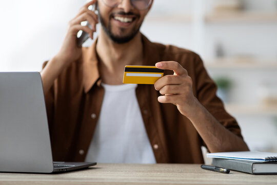 A man is engaged in a phone conversation while holding a credit card in his hand. He is seated at a desk with a laptop and a notebook. The atmosphere is relaxed and focused.