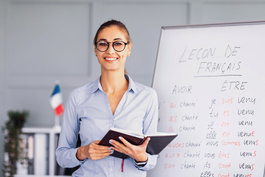 A cheerful teacher stands in front of a whiteboard with French grammar notes. She holds a notebook and smiles, encouraging students as a flag adds color to the setting.