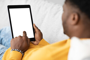 Modern digital tablet with blank screen in black guy hands, mockup. Unrecognizable african american man reclining on couch, using pad, scrolling, using newest mobile application, cropped