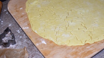 Raw cookie dough rolled out on a wooden board with Christmas tree shapes cut out using a metal cookie cutter. A festive home baking scene showing pastry preparation for holiday cookies.