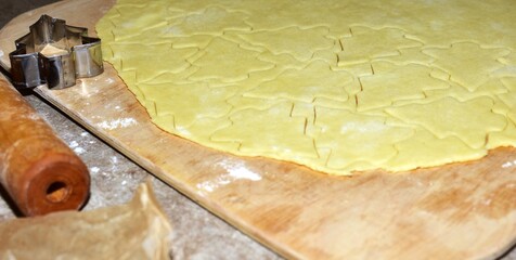 Cookie dough rolled flat on a wooden board, cut into Christmas tree shapes. A metal cutter and rolling pin lie nearby, showing the process of preparing holiday cookies at home.