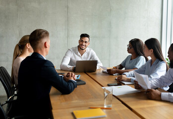 In a bright office setting, a diverse group of young professionals engages in a collaborative meeting. They brainstorm new project ideas while the Middle Eastern leader listens attentively.