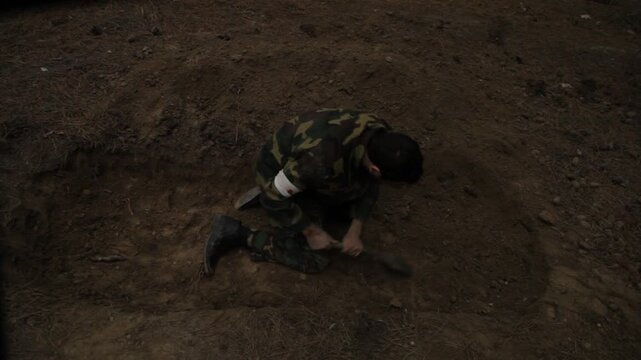 An exhausted soldier in camouflage buries a fallen comrade in a forest trench. He frantically covers the body with dirt using a shovel, surrounded by silent tall pine trees.

