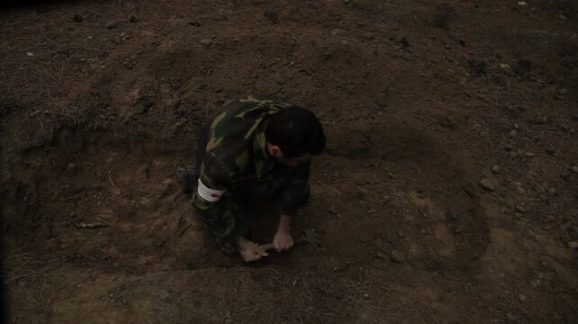 An exhausted soldier in camouflage buries a fallen comrade in a forest trench. He frantically covers the body with dirt using a shovel, surrounded by silent tall pine trees.

