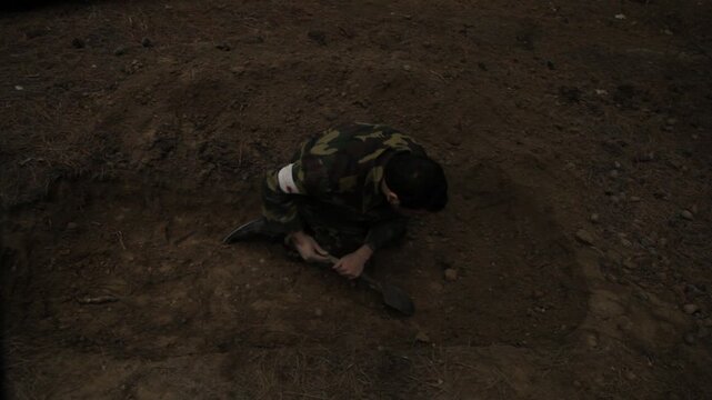 An exhausted soldier in camouflage buries a fallen comrade in a forest trench. He frantically covers the body with dirt using a shovel, surrounded by silent tall pine trees.

