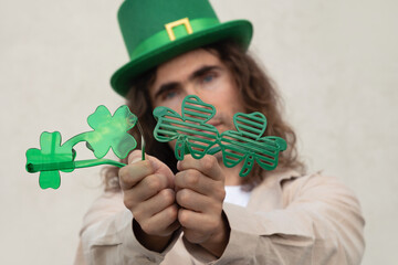 Young man choice funny Shamrock clover glasses wearing Green St. Patrick's Hat against beige wall. Close up.