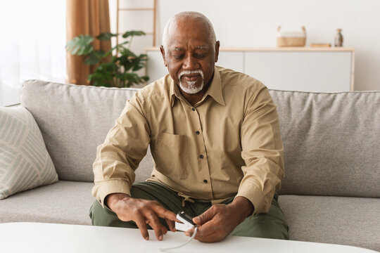 A senior man with gray hair is focused on a device while sitting on a couch in a warm and inviting living room. Sunlight filters in through the window, illuminating the space. - Powered by Adobe