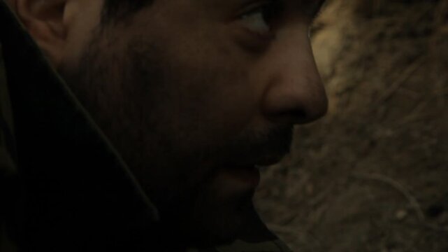 Close-up of nervous bearded soldier hiding in earthen trench. He smokes cigarette with shaking hands, eyes darting fearfully. He glances at silent walkie-talkie on the ground.