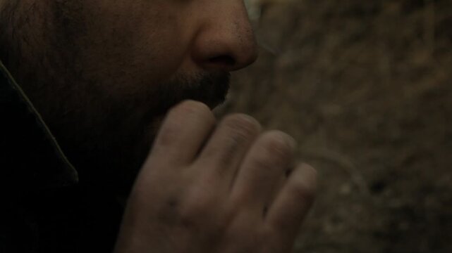 Close-up of nervous bearded soldier hiding in earthen trench. He smokes cigarette with shaking hands, eyes darting fearfully. He glances at silent walkie-talkie on the ground.