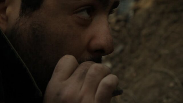 Close-up of nervous bearded soldier hiding in earthen trench. He smokes cigarette with shaking hands, eyes darting fearfully. He glances at silent walkie-talkie on the ground.