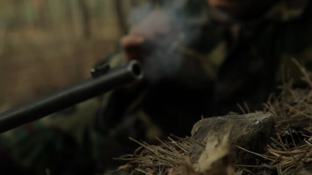 Close-up of camouflaged soldier placing lit cigarette on rock next to rifle barrel. He leaves it smoking on the stone to grip his weapon with both hands.