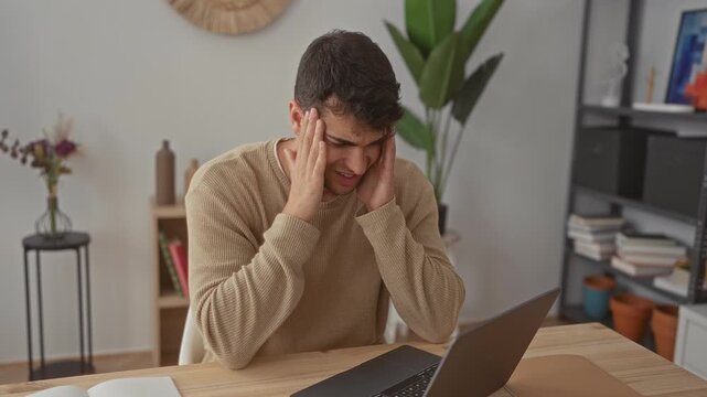 Man rubs head while holding laptop in hispanic home showing frustrated stress as young guy leans forward with a pained expression.