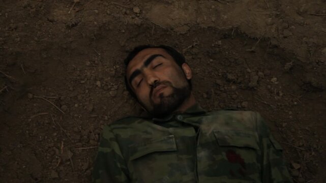 Top view of a dead soldier falling into a grave. The man in camouflage uniform has been shot and lies lifeless with wide open eyes on the dirt.